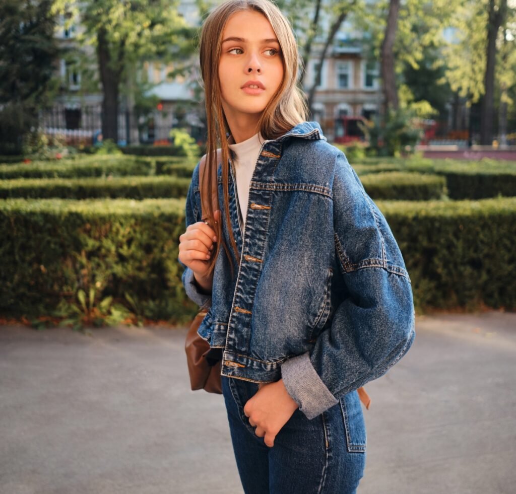 A young woman wearing a blue denim bomber jacket with rolled cuffs, standing outdoors in a park setting, carrying a brown bag.
