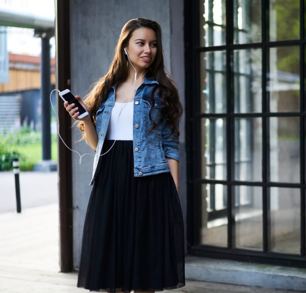 “A young woman stands outdoors wearing a light blue denim bomber jacket over a white top and a black midi skirt. She has long wavy hair, white earphones, and is holding a smartphone with a cord connected to her ears. She’s standing near a modern glass windowed building with a wooden pillar and a blurred green background.”