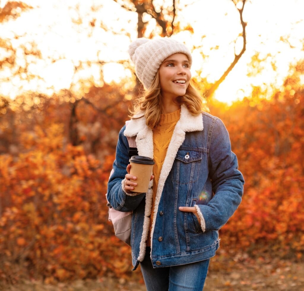 A young woman walking outdoors in autumn leaves, wearing a blue denim bomber jacket with a sherpa lining, a knit beanie, and jeans. She holds a hot coffee cup and smiles, the golden light suggesting a chilly, sunny day.
