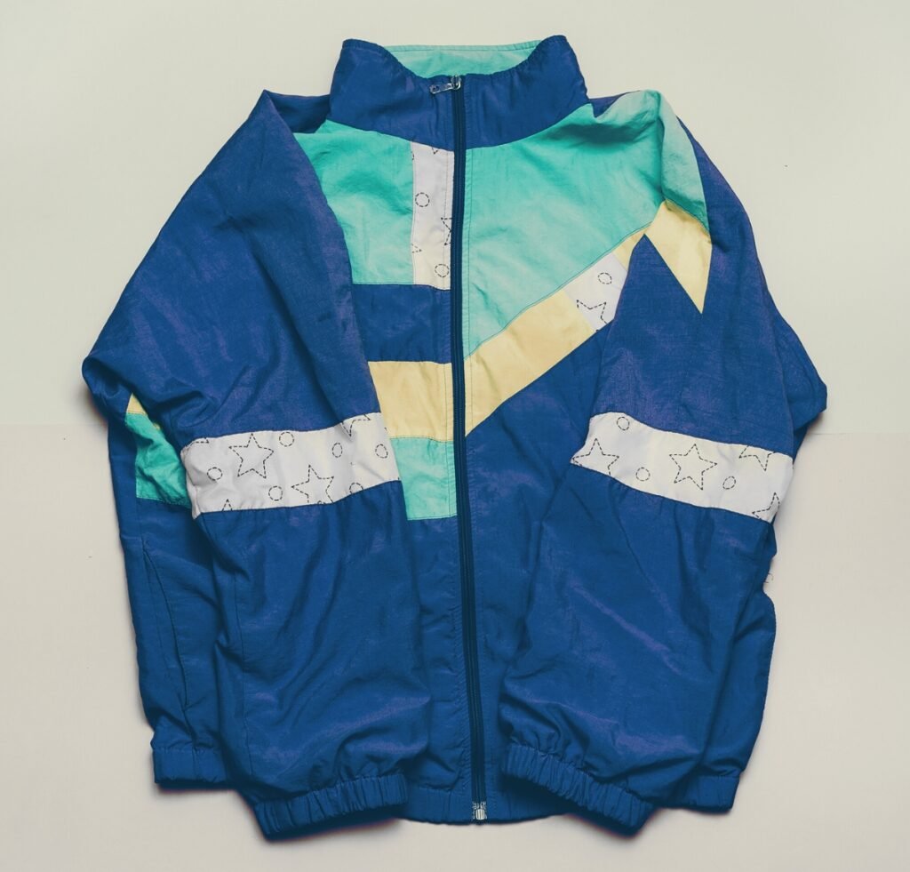 “Nylon bomber jacket with a color-block design in blue, teal, seafoam, and pale yellow, featuring diagonal and horizontal color panels, a front zipper, elastic waistband and cuffs, and decorative star-patterned white panels.”
