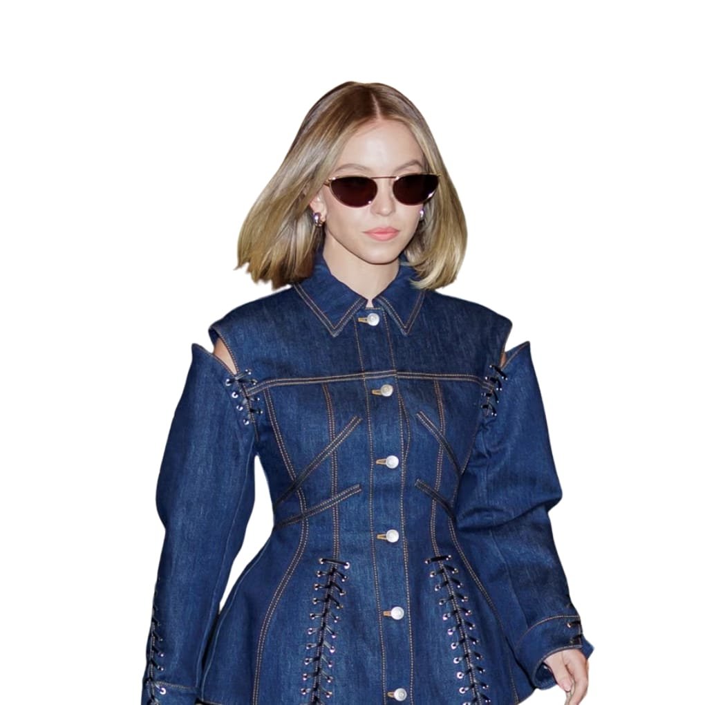 A woman wearing a dark blue denim jacket with corset-inspired lacing along the sides and shoulders, oversized puff sleeves with cut-out shoulders, silver buttons, and dark sunglasses, standing against a white background.