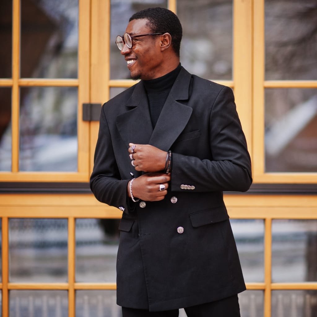 A stylish man wearing a black double-breasted overcoat, sunglasses, and a black turtleneck. He stands in front of a wooden-framed window, posing with his hands adjusting the coat and smiling