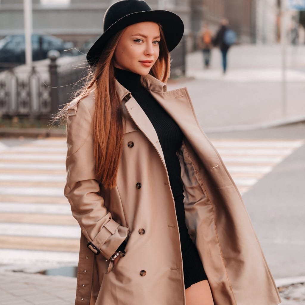 A stylish young woman wearing a beige double-breasted trench coat over a black knit top, black wide-brim hat, and has long straight hair. She stands on a city sidewalk near a crosswalk, with hands in coat pockets and a confident expression
