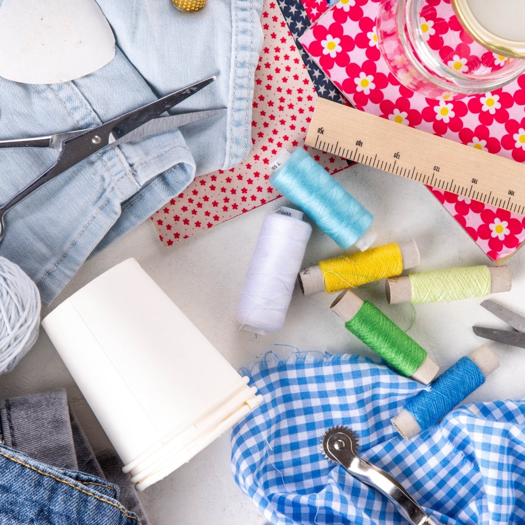 A craft workspace with cords and fabrics: corduroy and wool textiles, sewing tools, colorful thread spools, scissors, a ruler, and patterned fabrics on a light surface.