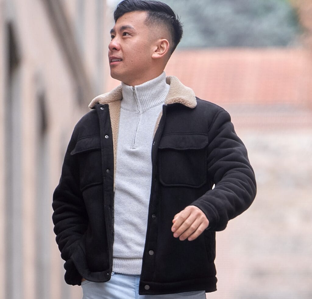 A man wearing a black B-3 bomber jacket with a beige shearling collar over a light gray zip-neck sweater, standing outdoors with one hand in his pocket and looking to the side.