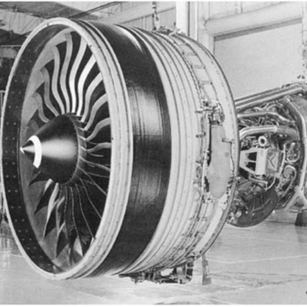 A large jet engine nacelle with a multi-blade turbine and connected mechanical systems in a hangar.