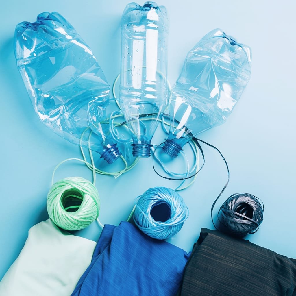 Assorted blue and teal plastic bottles and plastic bags arranged around spools of colorful yarn and folded fabric pieces on a light blue background.