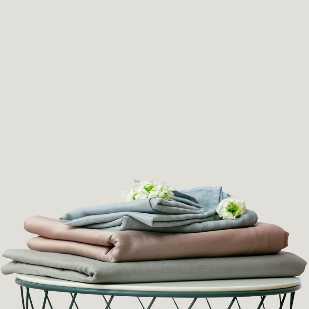 Stack of neatly folded pastel-colored bedsheets in cream, blush, and blue tones on a round table, with soft white flowers resting on top, set against a light gray background.