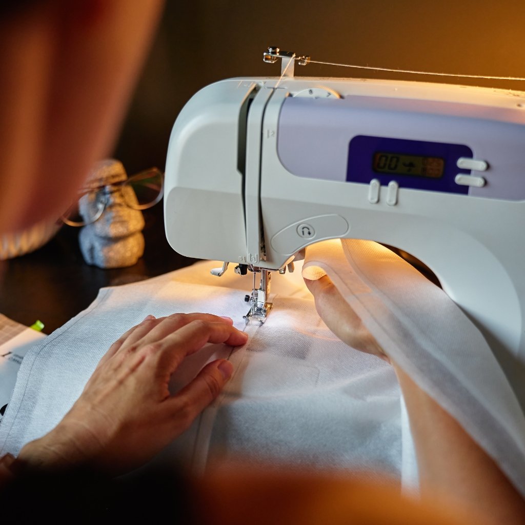A person guiding white fabric under a sewing machine needle as it stitches; a hand rests on the fabric while the performer’s other arm guides the material.

