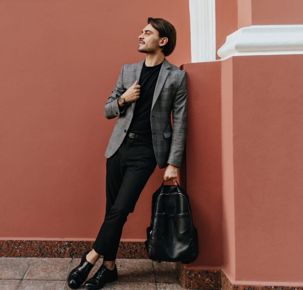 A stylish man leans against a warm terracotta wall, wearing a grey checkered blazer, black shirt and trousers, black loafers, and carrying a black backpack. Butcher’s Linen Fabric