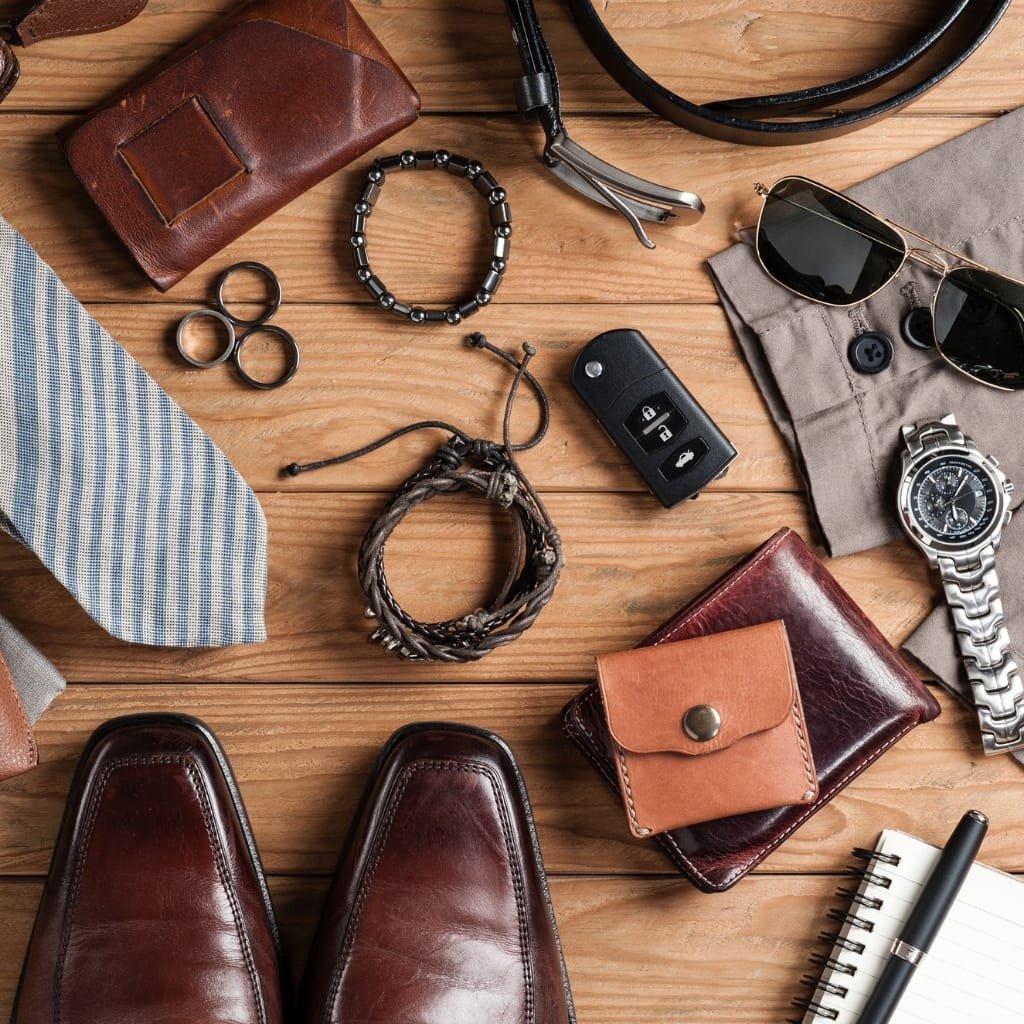 A flat lay of men’s accessories on a wooden surface, including brown leather shoes, a striped tie, bracelets, a car key fob, sunglasses, a watch, rings, belts, wallets, a notepad with a pen, and a small brown leather pouch.