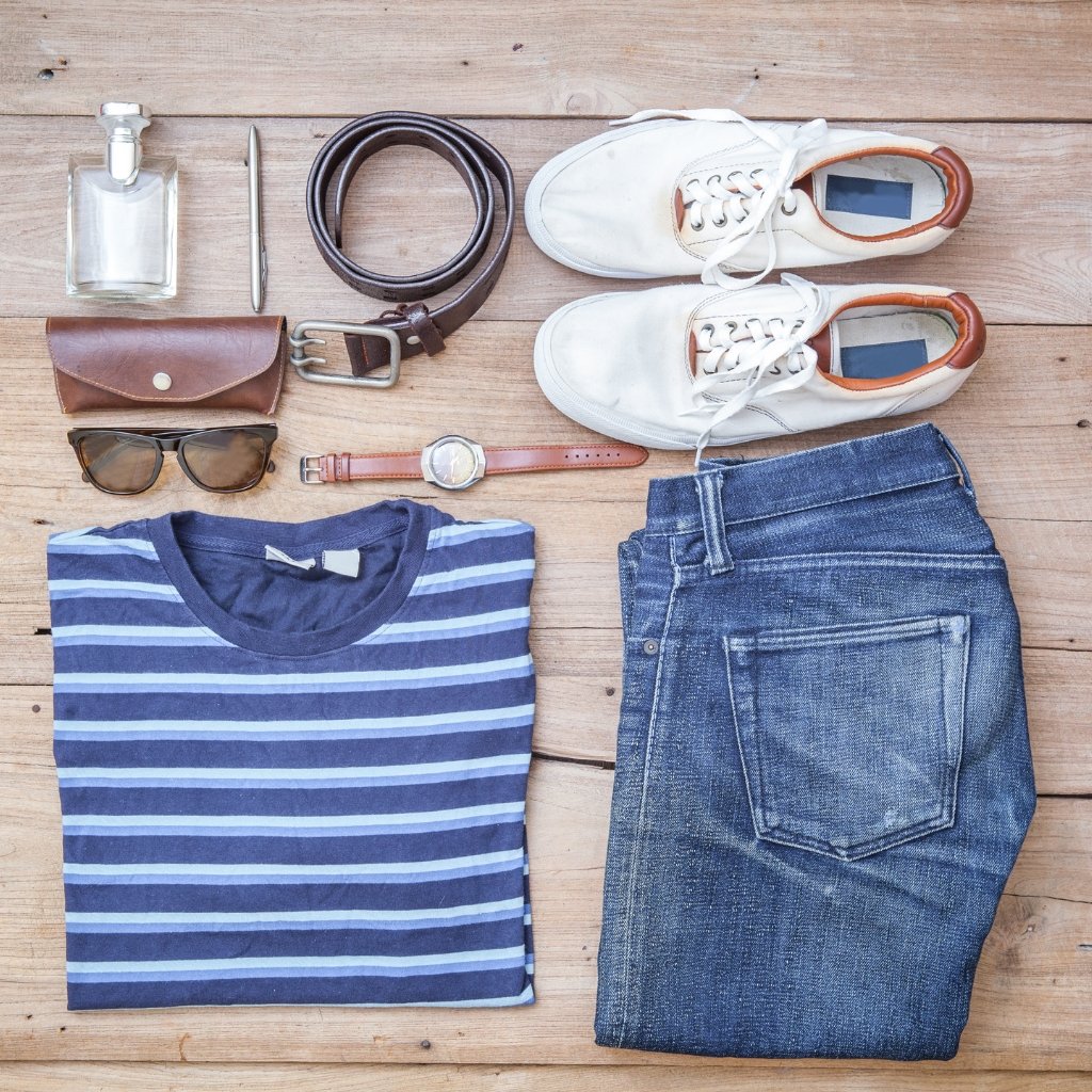 A flat lay of a casual outfit on a wooden surface, including a striped navy angora wool shirt, denim jeans, white sneakers, brown leather belt and accessories, and a bottle of cologne.