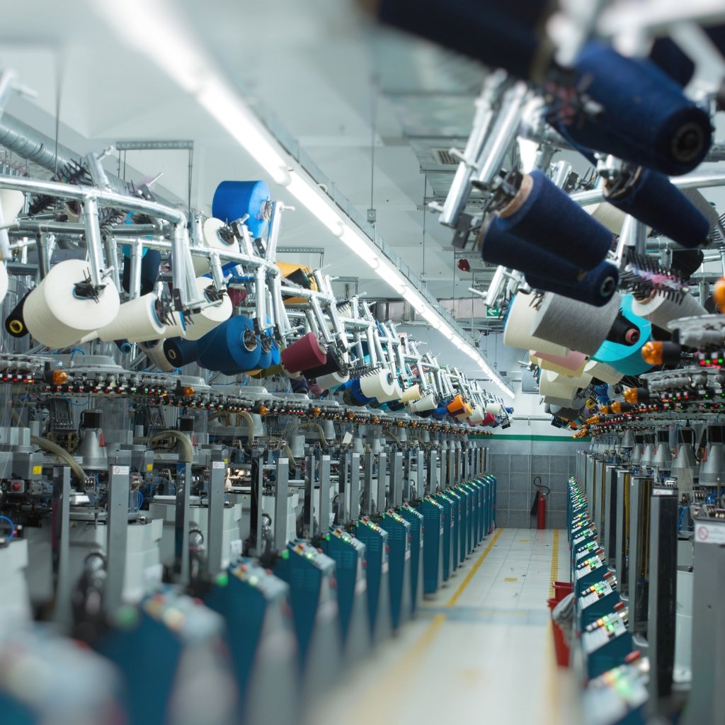 Industrial textile manufacturing line with multiple spools and fabric rolls in various colors, suspended above machinery along a long row.