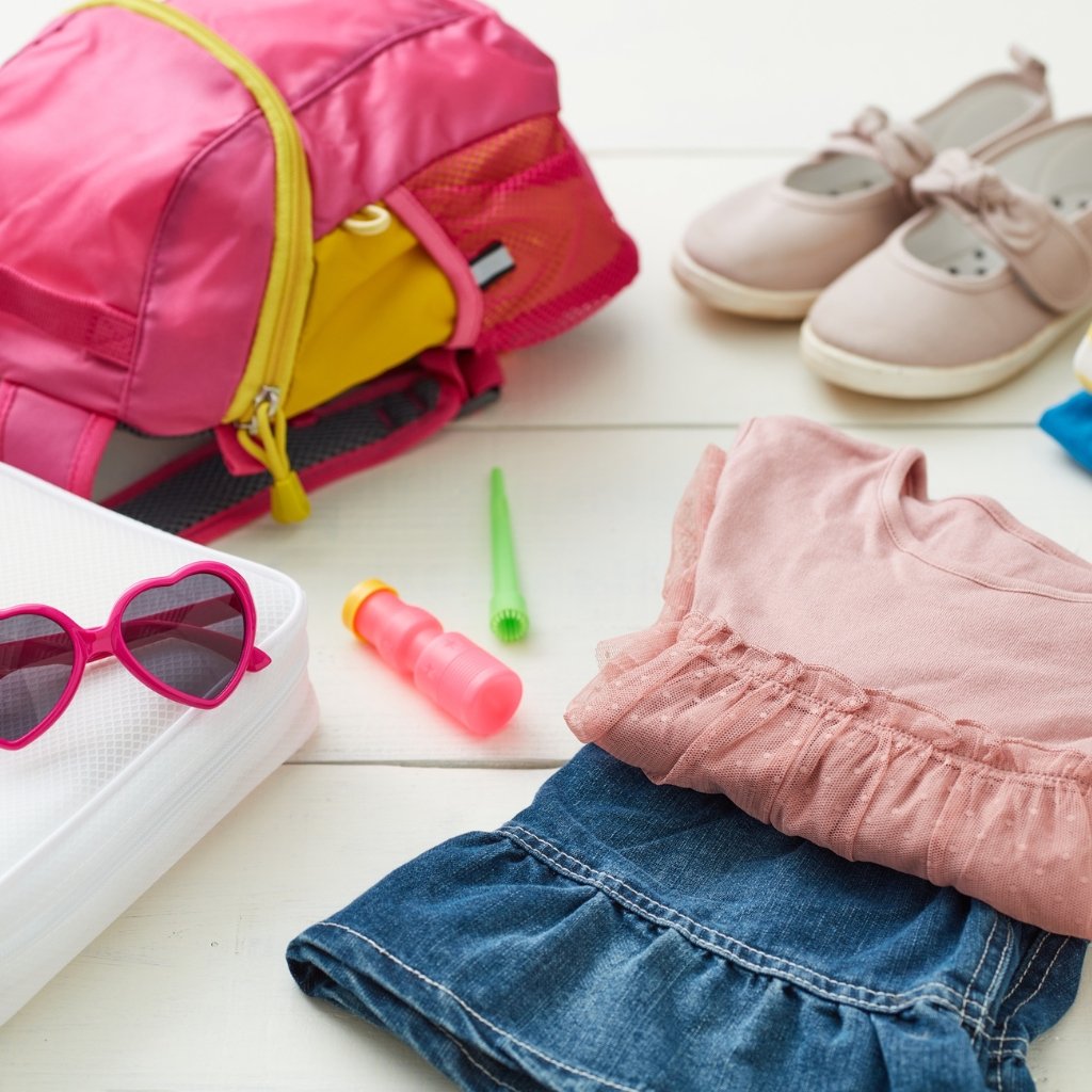A neatly arranged travel scene featuring a pink backpack, pairs of pink sunglasses, a pink top, folded denim shorts, and small travel items on a light wooden surface; tweed wool fabric is not prominently visible in this image.