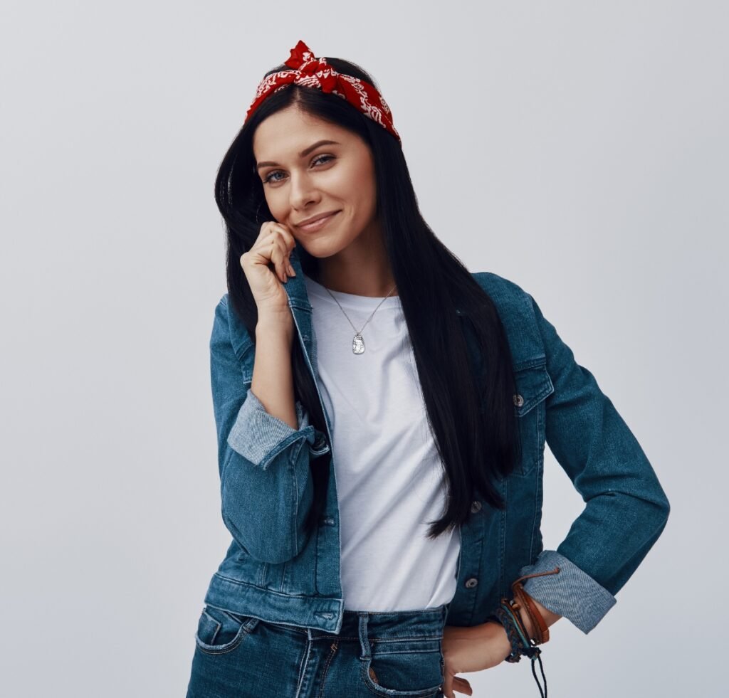 A young woman with long dark hair wearing a red bandana headband, a white t‑shirt, and a blue denim jacket over denim jeans. She is smiling softly and resting her chin on her hand against a light gray backdrop.