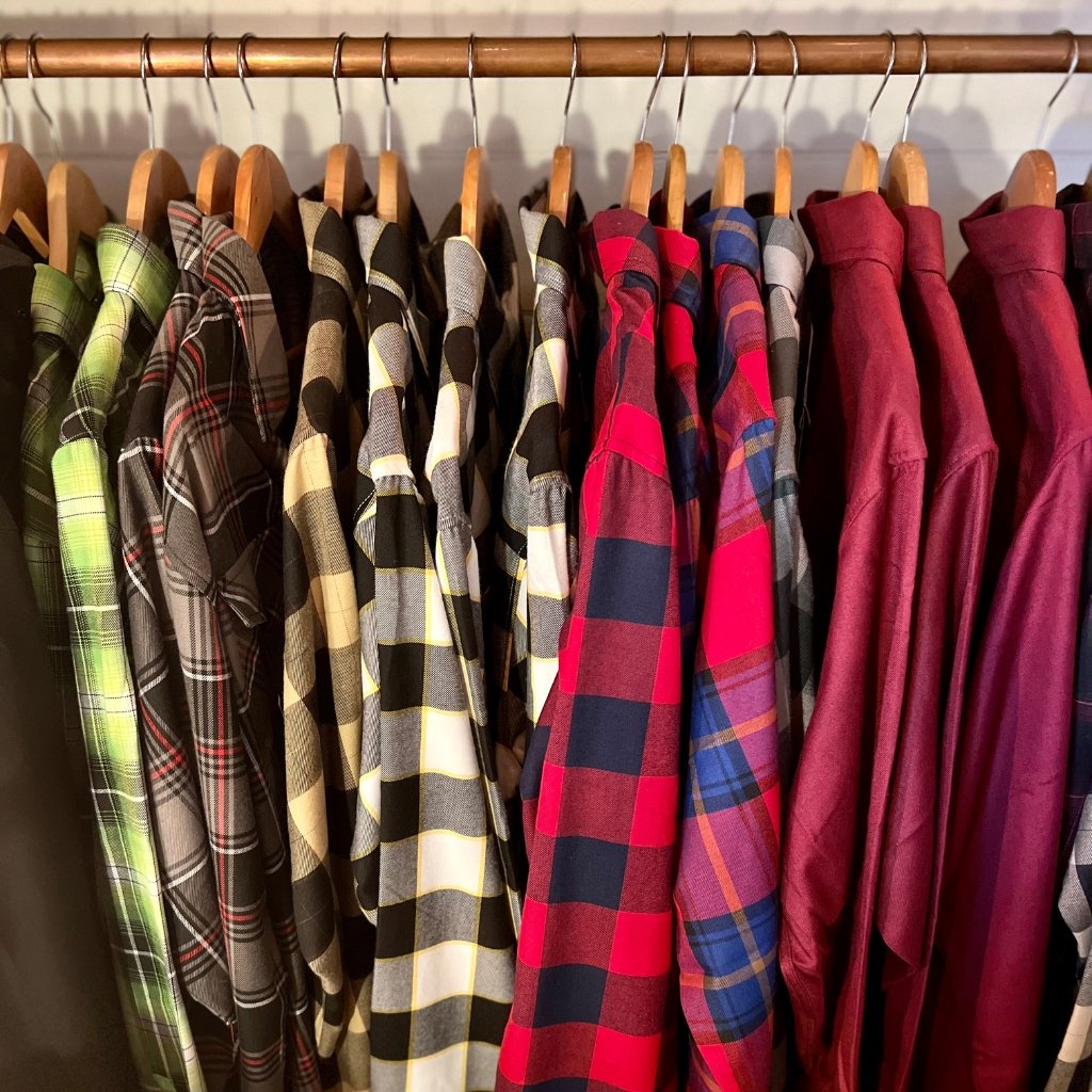 A row of plaid/flannel shirts hanging on wooden hangers in a closet, featuring various colors including greens, blacks, creams, reds, and blues, with soft, cozy textures.