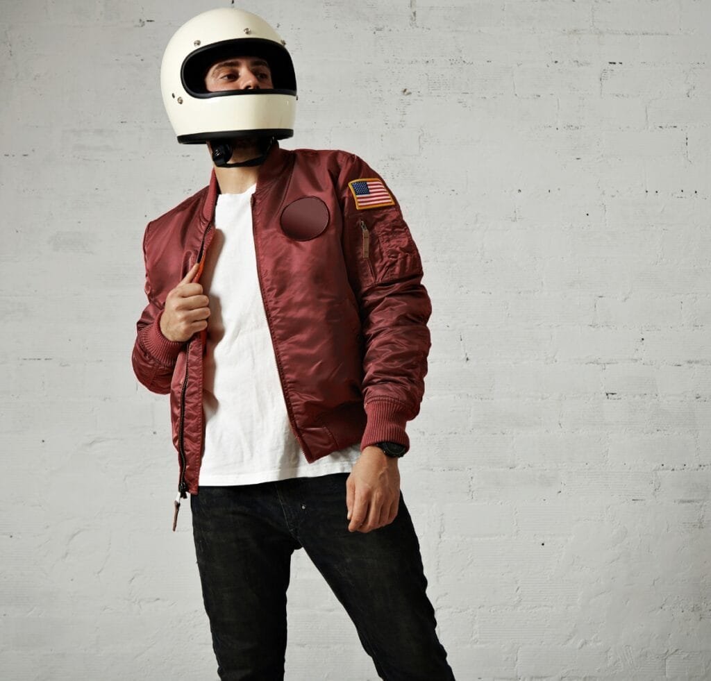 A person wearing a white helmet and a maroon flight bomber jacket, standing against a light gray brick wall. The jacket has an American flag patch on the sleeve, a circular patch on the chest, and a zipper pocket on the sleeve. The person is pulling the jacket slightly open over a white T‑shirt, wearing black jeans and a wristwatch. Types of Bomber Jackets