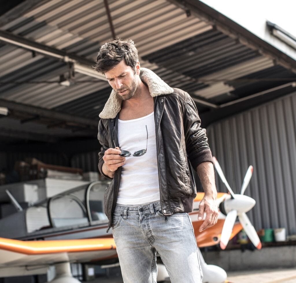A stylish man stands in a hangar next to a small orange-and-white aircraft. He wears a brown bomber jacket with a shearling-lined collar over a white tank top and light-washed jeans. He holds a pair of sunglasses in one hand, looking down with a relaxed, confident pose.