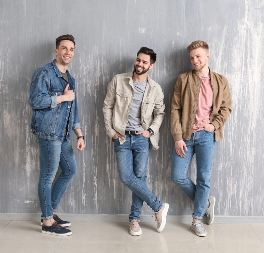 “Three men leaning against a gray textured wall, wearing casual bomber jackets and jeans, smiling at the camera.” The History of Bomber Jackets