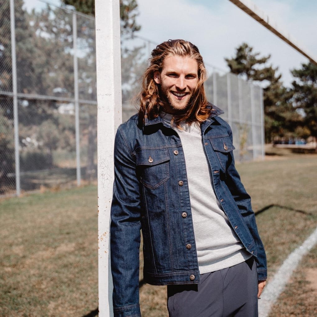 A man with long blond hair and a denim jacket leans against a white post on a sunny day at a sports field, smiling at the camera.