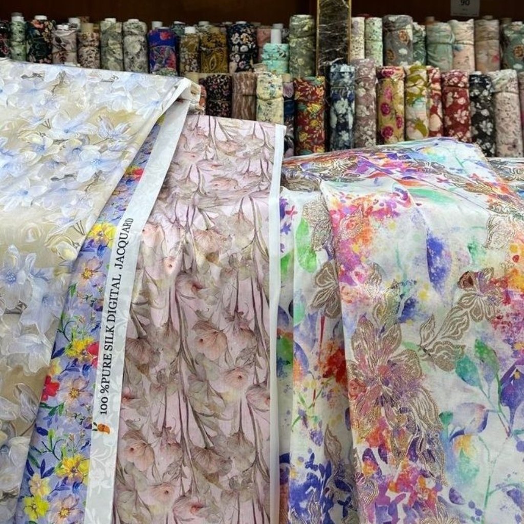 A display of rolled fabrics in a shop: sheer printed organza with delicate floral patterns in pale pinks, whites, and soft colors. Behind them are stacked rolls of colorful, patterned fabrics, including metallic and botanical designs.