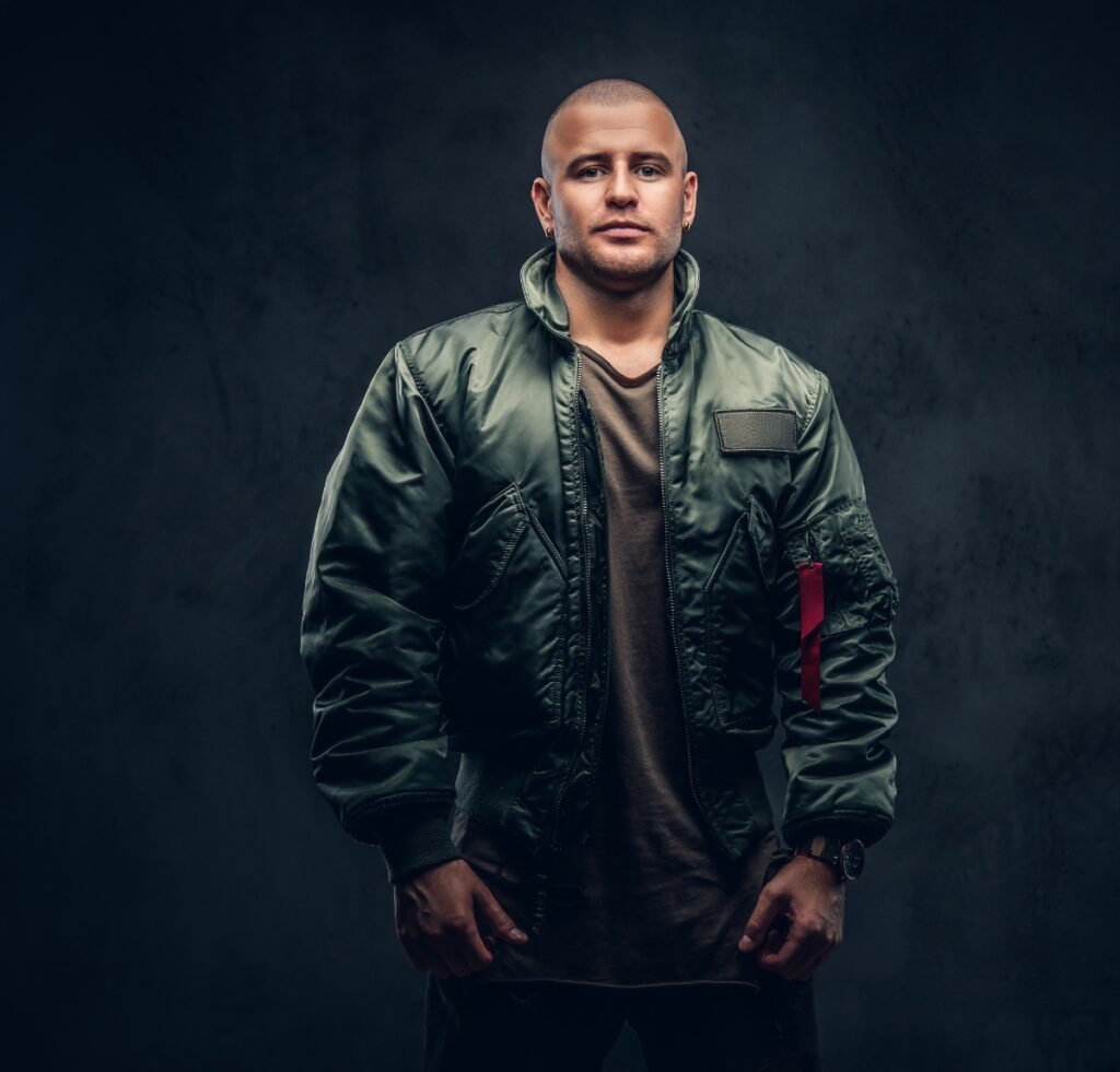 "Portrait of a man wearing a green bomber jacket over a brown shirt, standing against a dark textured background. He has a short haircut and a calm, confident expression." Bomber Jacket in Nylon