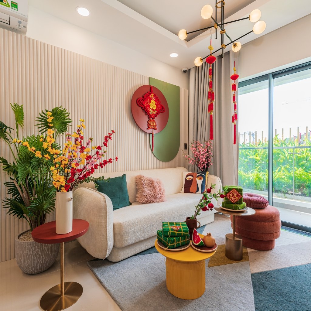 A bright, modern living room with a white velvet sofa adorned with teal and pink cushions. A round yellow coffee table holds decorative cakes and fruit, and a red ottoman sits beside a tall vase of yellow and red flowering branches. Large windows show green plants outside, with festive red hanging decorations from the ceiling and a textured wall behind the sofa.