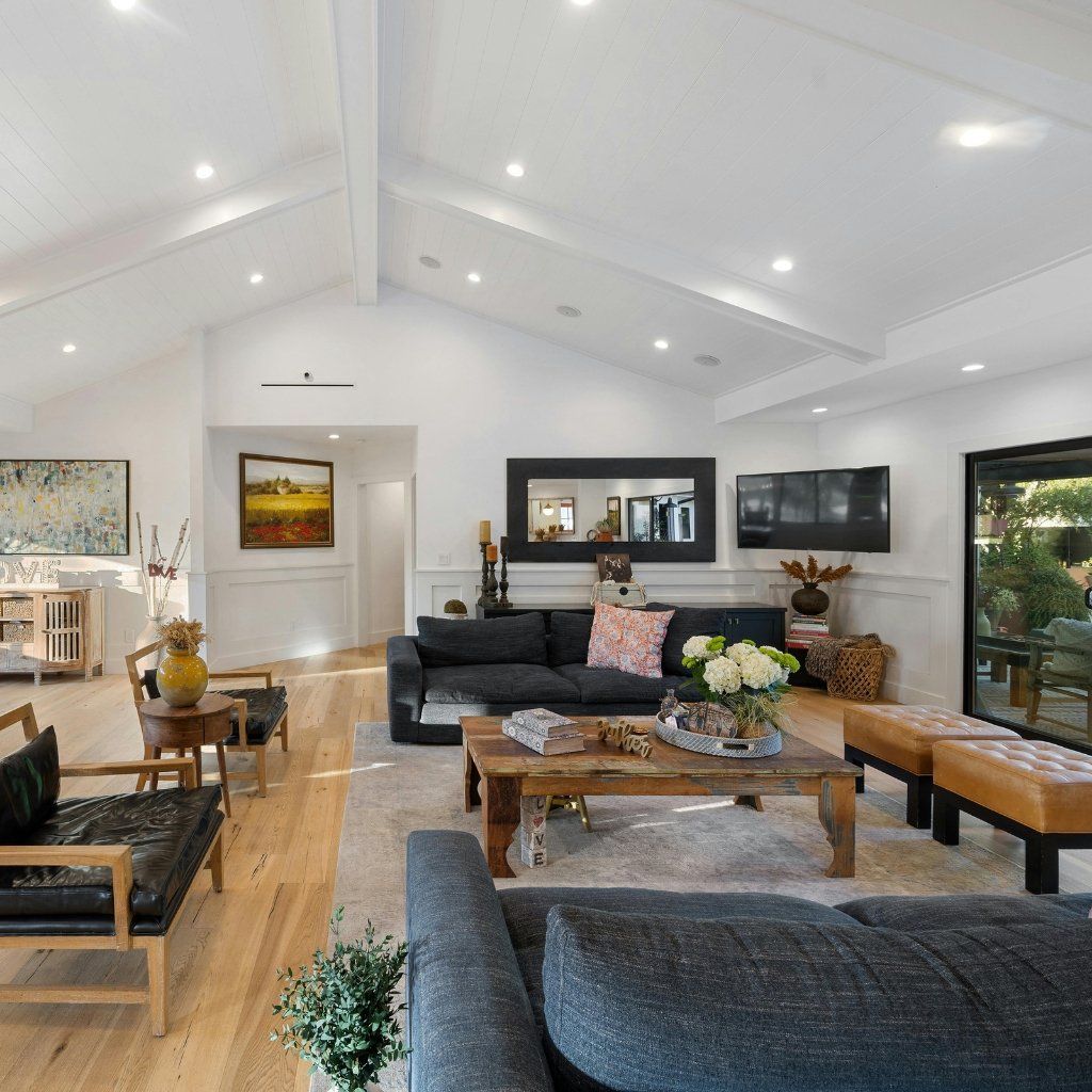 A bright, airy living room with white walls and vaulted white paneled ceiling. A large navy sofa and matching chairs surround a rustic wooden coffee table. A wall-mounted flat-screen TV and a long dark console with decorative items sit against the back wall. Light hardwood floors, a light-colored area rug, and two tan leather ottomans add warmth. Artwork and plants decorate the space, with natural light streaming in from a glass door to the right.
