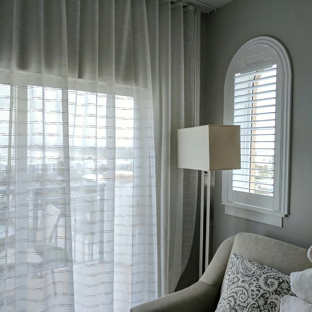 A bright, airy living room corner featuring sheer organza curtains draped over a large window, with soft natural light filtering through. A tall floor lamp stands near a beige upholstered sofa with decorative patterned cushions, and a white arched window with shutter slats is mounted on the wall behind.