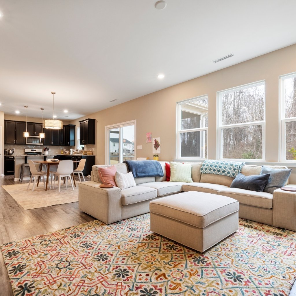 A bright, open-concept living room with a large beige sectional, colorful throw pillows, and a patterned rug. Nearby, a modern kitchen with dark cabinetry and a wooden dining table. Large windows let in natural light, and a neutral color palette creates a cozy, airy vibe.