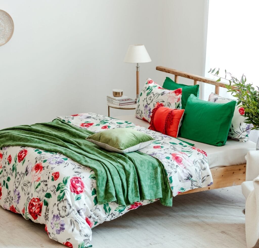A bright, stylish bedroom featuring a wooden bed dressed in floral bedding with green accents: green throw, green and red pillows, and a matching floral duvet. A side table with a lamp sits beside the bed, and a plant decorates the headboard area. Hebei Linen Fabric