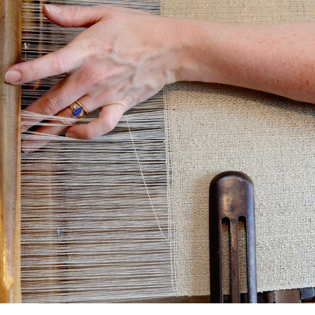 A close-up photo of a person’s hand operating a traditional handloom. The fingers guide fine white threads across the loom, with a wooden loom beam on the left and a dark shuttle/press tool on the right. The hand wears a ring with a blue stone