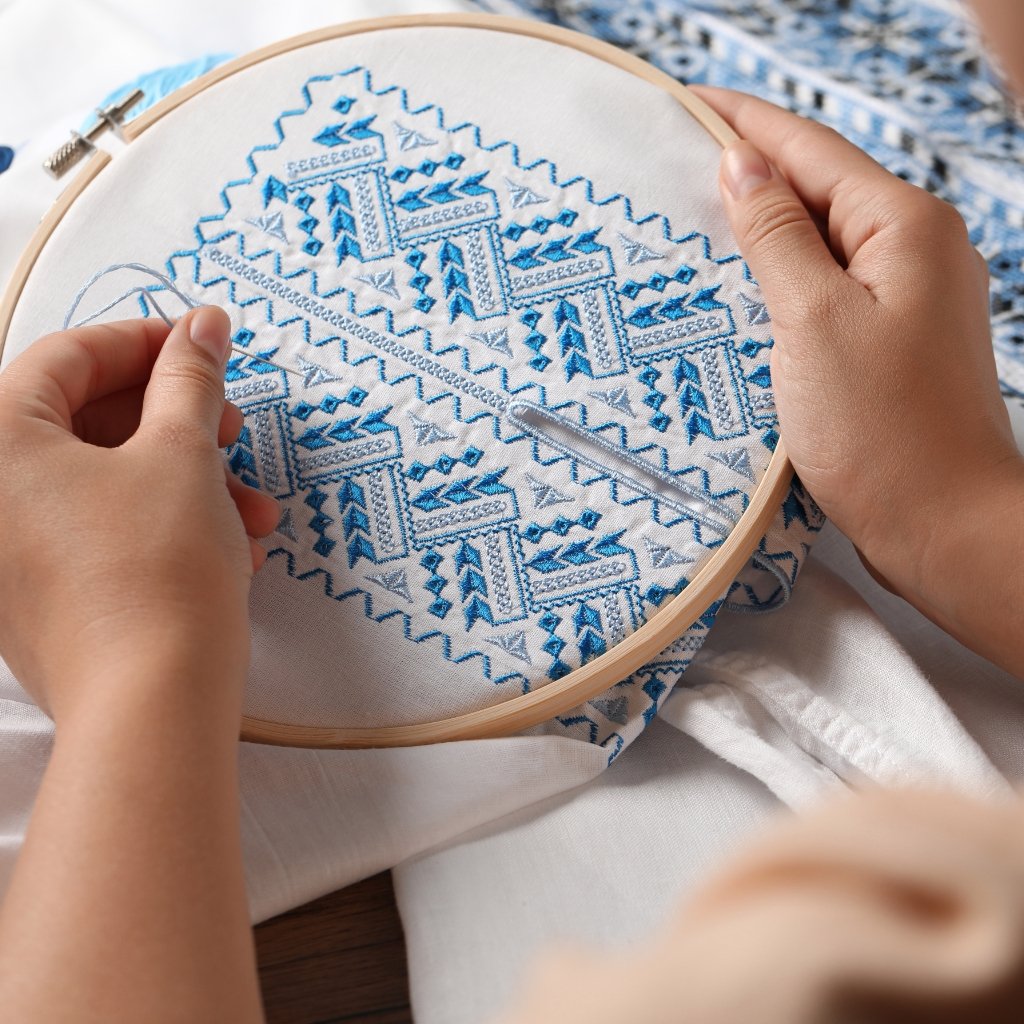 A close-up photo of a person embroidery hooping a white fabric with blue geometric embroidery patterns. The person’s hands guide a needle with light blue thread, creating zigzag and diamond motifs. The wooden hoop sits on a light surface, with more stitched fabric visible in the background.