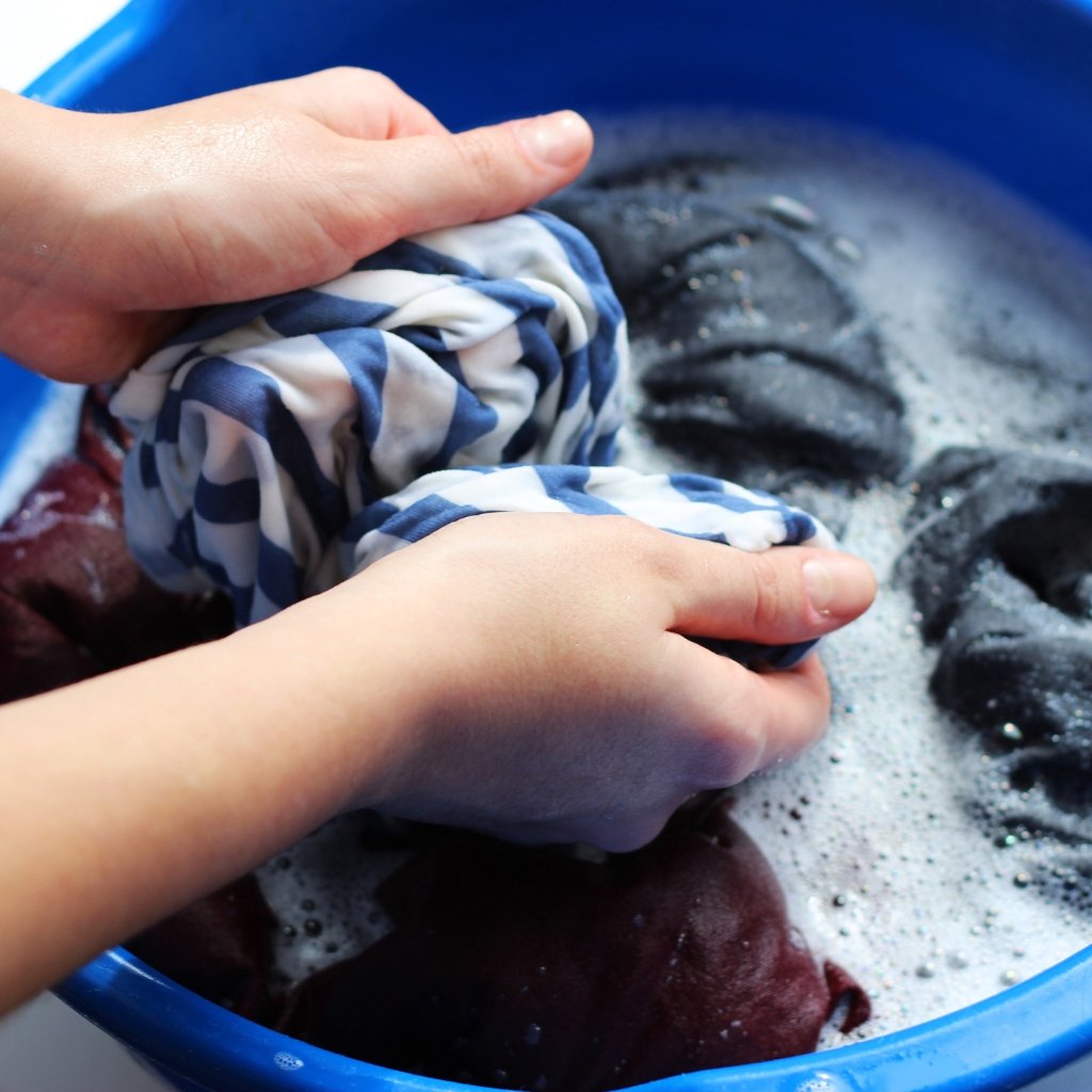 A pair of hands gently wringing a delicate, translucent organza fabric in a blue basin filled with soapy water.
