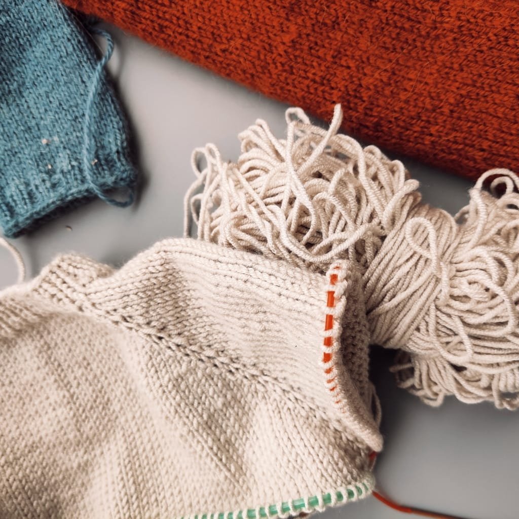 A close-up view of assorted knitted fabrics and yarns. In the foreground is a cream-colored knitted textile with long fringe on the right; a bundle of tangled off-white yarn sits behind it. In the background, a teal knitted piece is on the left and a rust-orange fabric is at the top.