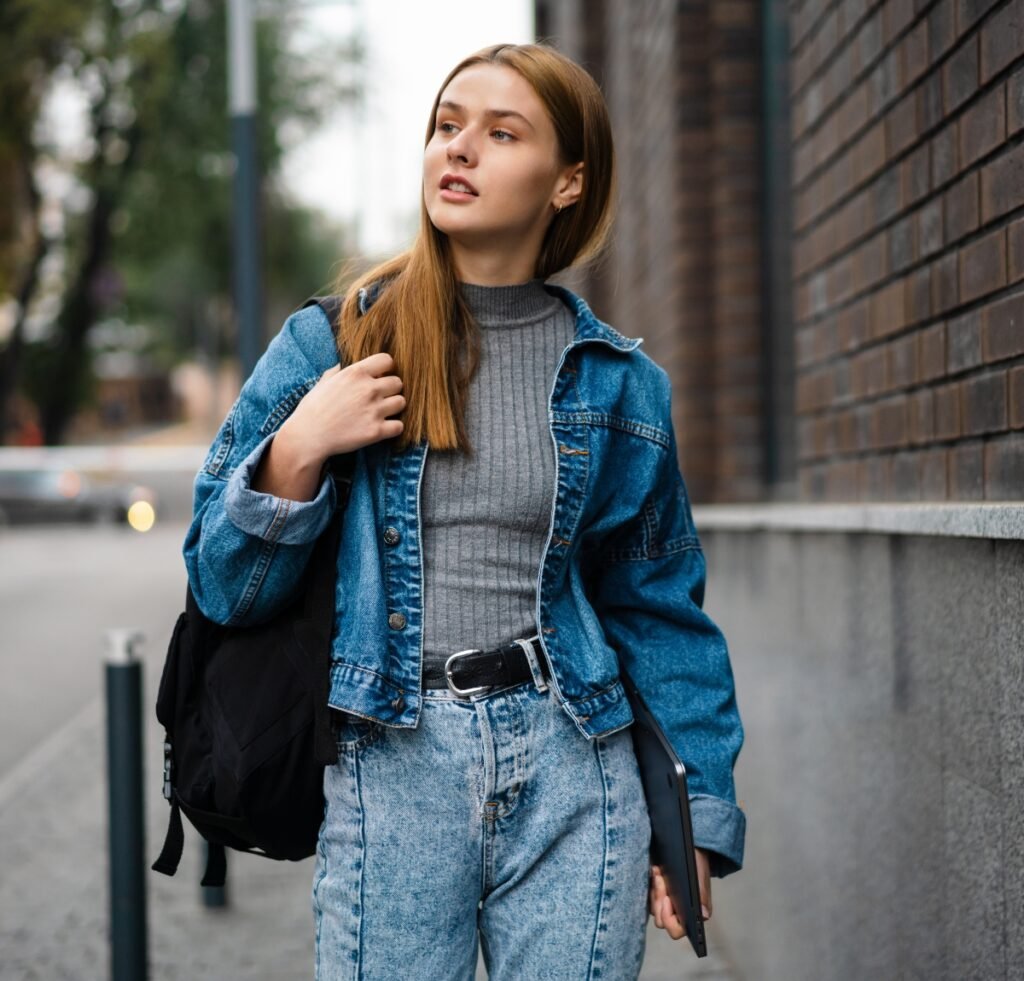 A young woman stands on a city sidewalk beside a brick wall. She wears a blue denim bomber jacket over a gray ribbed turtleneck, high-waisted light-wash jeans, and a black belt. She carries a black backpack on one shoulder and holds a tablet or notebook in her other hand. Her straight, light brown hair is parted to the side, and she gazes slightly upward with a confident expression.