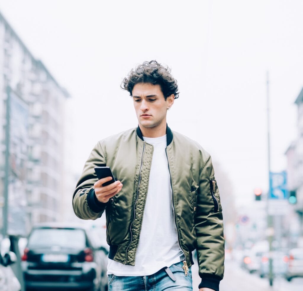Flight Bomber Jacket: Timeless Style Meets Modern Utility 4 A young man walks down a city street while looking at his smartphone. He wears a olive-green flight bomber jacket over a white T-shirt and light-wash jeans, with a focused expression. Urban buildings and parked cars blur in the background.
