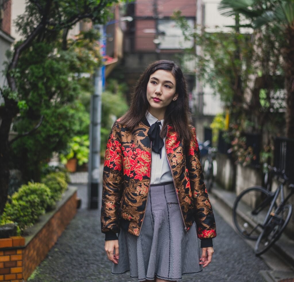 A stylish young woman stands on a quiet city street wearing a patterned bomber jacket with red floral designs, a white blouse with a black bow, and a grey pleated skirt.