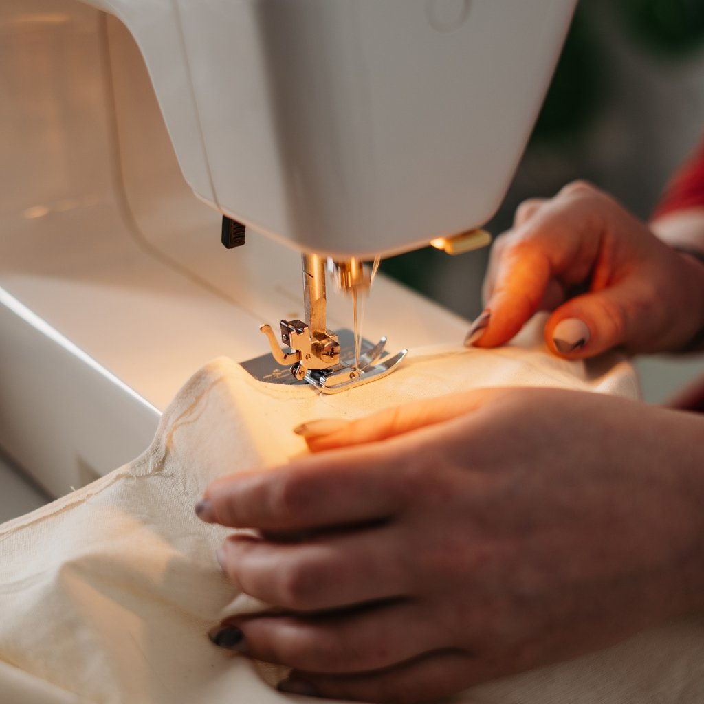  A close-up photo of a sewing machine stitching light, sheer fabric. The presser foot is down, and a pair of hands guides the fabric, creating delicate seam lines. The fabric appears to be crushed organza, with a subtle sheen and textured surface.