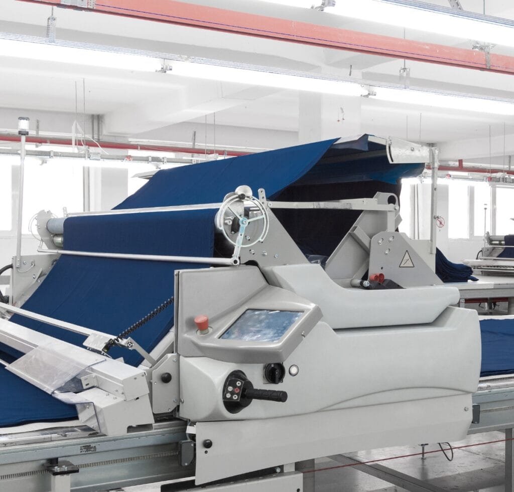 A modern textile production machine in a bright factory, weaving or finishing blue linen fabric off a conveyor with a large blue web, control panel, and mechanical components.