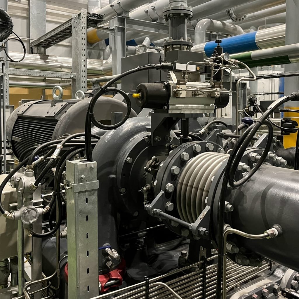 Industrial mechanical room with large turbine or pump, bolted pipework, hoses, and a compression/valve assembly mounted on a heavy steel frame.