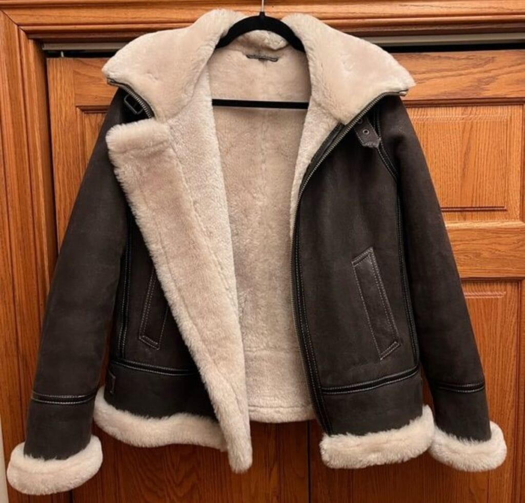 A dark brown shearling bomber jacket with white faux shearling lining and cuffs, hanging on a hanger. The coat is open, showing the soft white interior lining and large front pockets.