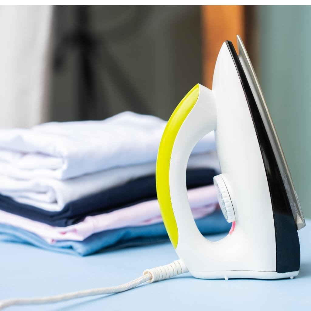 A modern electric iron standing on an ironing board with a stack of folded clothes in the background.
