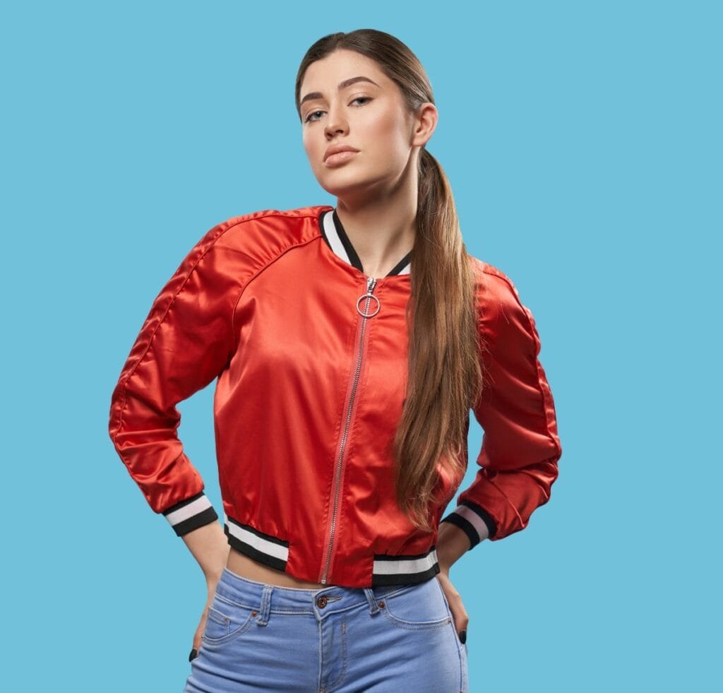 "A young woman wearing a shiny red satin bomber jacket with black and white striped cuffs and hem, standing against a solid blue background. She has long brown hair tied in a low ponytail and poses with hands in her pockets."