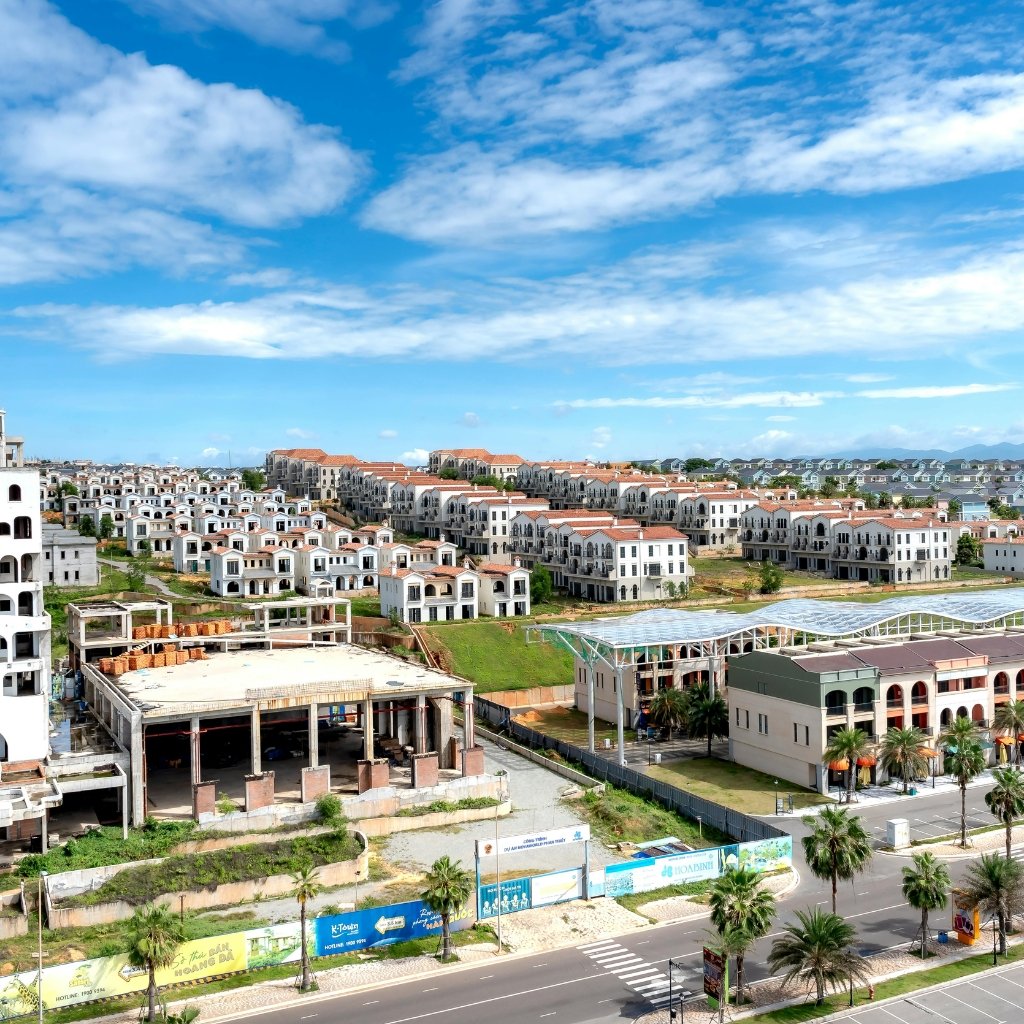 Why Supima Cotton Feels Softer, Stronger, and Smarter 23 A coastal residential development under construction: rows of white, Mediterranean-style houses with red-tiled roofs stretch across a bright, blue sky. A partially built multi-story structure sits in the foreground, with banners along the street and palm trees lining the road.