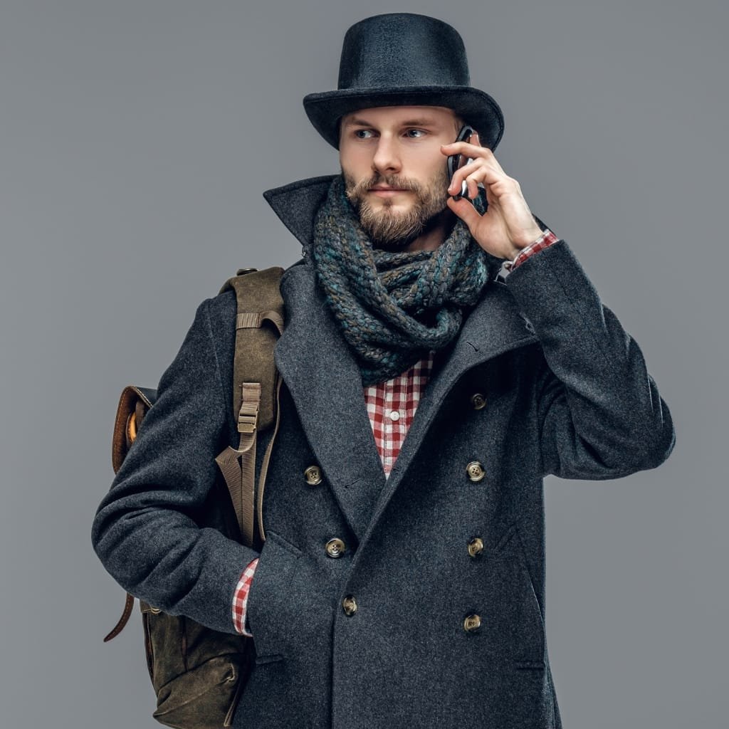 "Man wearing a dark double-breasted wool coat with a large collar, scarf, and hat, talking on a smartphone while carrying a brown backpack