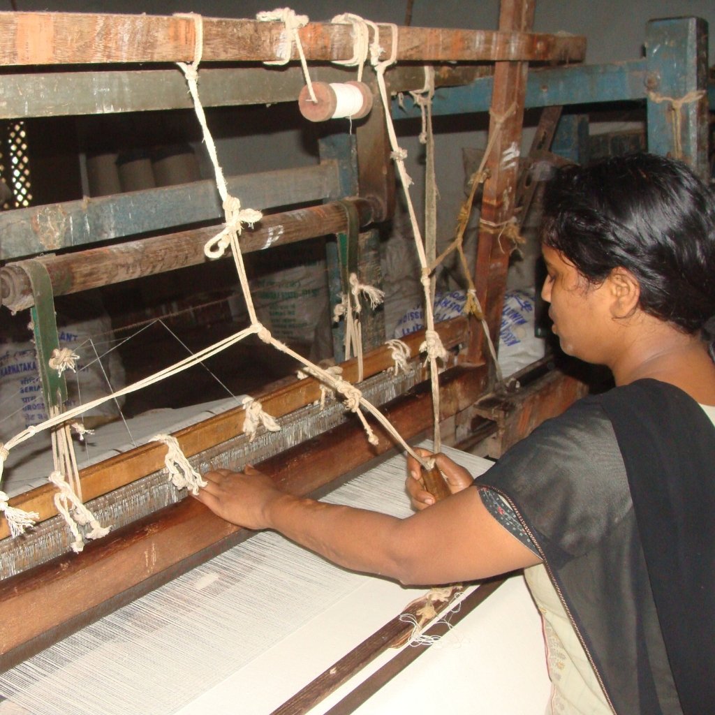 A artisan operates a traditional loom, weaving white khadi fabric by hand. Strings, wooden beams, and spinning components surround the worker as she guides the yarn across the loom.

