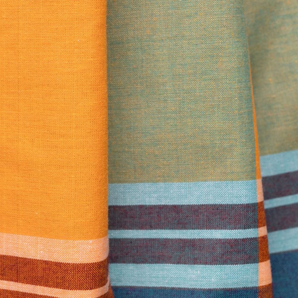 Close-up of several Khadi cotton fabrics in warm orange and teal hues, showing a coarse, handmade weave with visible texture and colored stripes at the bottom edge.

