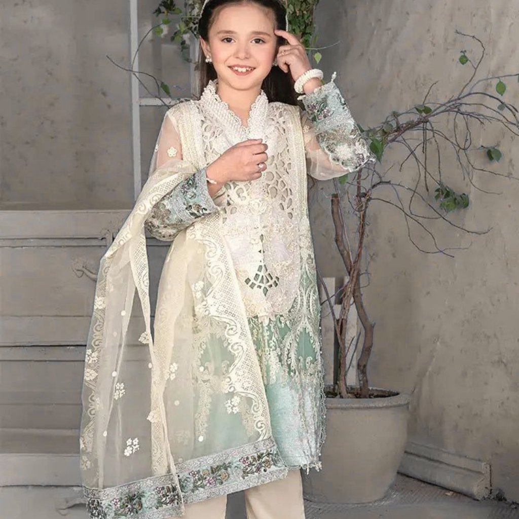 A smiling young girl wearing a delicate, cream-colored lace traditional outfit with sheer sleeves and an embroidered overlay. She stands indoors near a potted plant, posing with one hand near her head and the other resting on her chest.