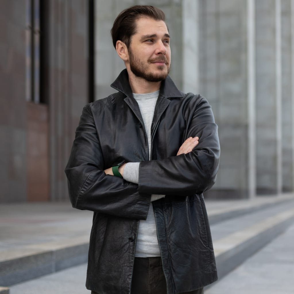  A man with a trimmed beard wearing a black leather coat, standing outdoors with arms crossed and looking to the right